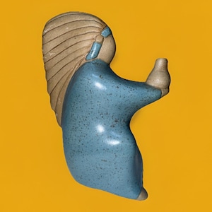 May include: A ceramic figurine of a person in a blue dress holding a small vase. The figurine has tan hair and a blue headband. The figure is set against a yellow background.