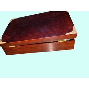 May include: A rectangular wooden box with a hinged lid. The box is a dark reddish-brown color with a glossy finish. The corners are reinforced with gold-colored metal. The box is open, revealing the interior.