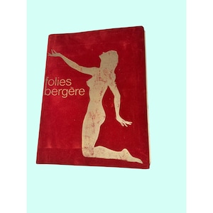 Puede incluir: Una portada de libro vintage con fondo rojo y silueta dorada de mujer arrodillada. Las palabras "folies bergère" están impresas en dorado en la portada.