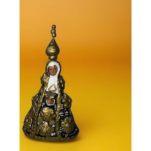 Op de afbeelding: Gedetailleerd beeldje van een religieuze figuur, met een goud- en zwart kleurenschema. De figuur is versierd met ingewikkelde bloempatronen en een hoge, sierlijke hoofdtooi. De achtergrond is effen warmgeel.