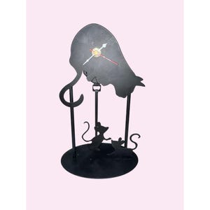 May include: Black metal clock with a unique design featuring a silhouette of a figure with a long tail and two mice at the base. The clock hands are white with a red second hand. The clock is on a round base.