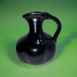 May include: A small, black ceramic pitcher with a rounded body and a curved handle. The pitcher has a glossy finish.