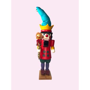 May include: A wooden nutcracker in the shape of a clown. The clown has a red coat with green and purple pants. It has a blue hat with a yellow crown and is holding a golden staff. A small doll is on its shoulder.