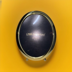 May include: Gold oval picture frame with a black velvet mat and a gold stand. The frame has the text "CARR International Photo Products" and "SOLID GLASS 8x10" printed on the mat.