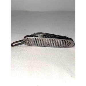 May include: A silver-toned folding pocket knife featuring a textured, cross-hatched metal handle and a small metal loop attached to one end. The knife is shown in a closed position with the blade tucked inside the handle, displaying a metallic finish.