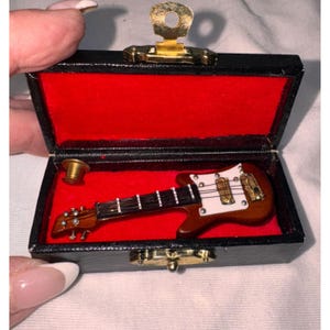 Puede incluir: Miniatura de guitarra eléctrica marrón dentro de un estuche negro con interior rojo. El estuche está abierto, revelando la guitarra y un pequeño objeto dorado. La guitarra tiene un golpeador blanco y herrajes dorados.