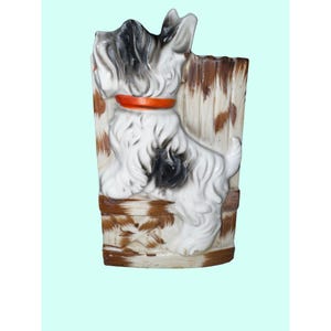 May include: A ceramic planter shaped like a Scottish Terrier dog. The dog is white with black and gray markings, wearing an orange collar. The planter has a brown and white wood-like background.