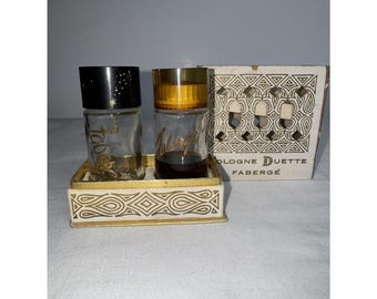 Puede incluir: Un juego Faberge Cologne Duette vintage. El juego incluye dos frascos de perfume de vidrio en una bandeja decorativa con detalles dorados. Una botella tiene una tapa negra con estrellas doradas, la otra tiene una tapa dorada. La caja es de color crema con un diseño geométrico.