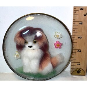 May include: A round glass paperweight containing a fuzzy, white and brown dog figurine with black eyes, positioned next to a wooden ruler showing a diameter of approximately 3.5 inches. The dog is surrounded by three small, colorful plastic flowers.
