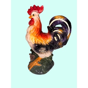 Puede incluir: Figura de gallo de cerámica colorida. El gallo tiene una cresta y una barbilla rojas, con plumas naranjas, amarillas y negras. Está posado sobre una base verde. La figura mide aproximadamente 30 cm de altura.