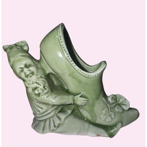 May include: A green ceramic figurine of a gnome holding a large shoe. The gnome is smiling and wearing a hat. The shoe has a clover design and a dotted trim. The figurine is a decorative item.