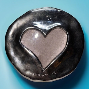 May include: A silver-toned, heart-shaped picture frame with a clear glass insert. The frame has a textured, organic edge and sits on a light blue surface. The words "Barefoot Dreams" are etched on the bottom.