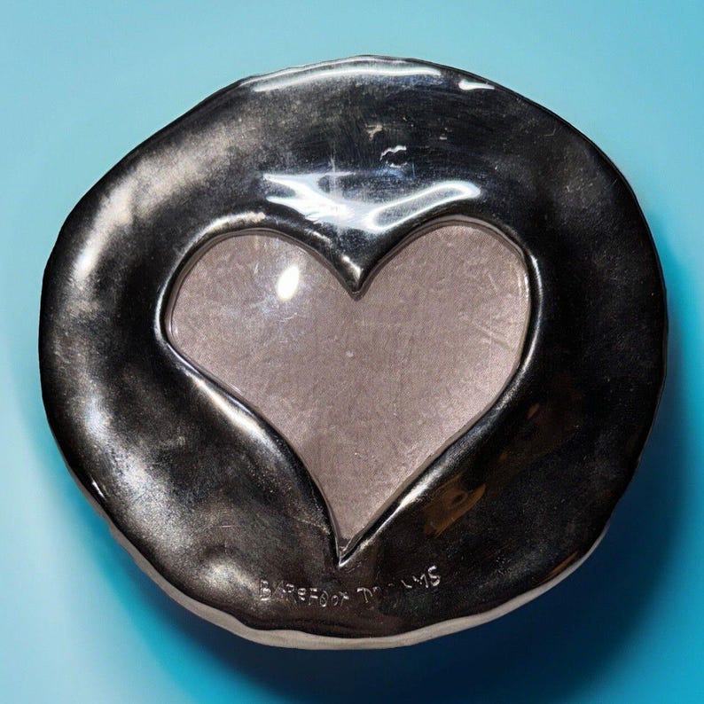 May include: A silver-toned, heart-shaped picture frame with a clear glass insert. The frame has a textured, organic edge and sits on a light blue surface. The words "Barefoot Dreams" are etched on the bottom.