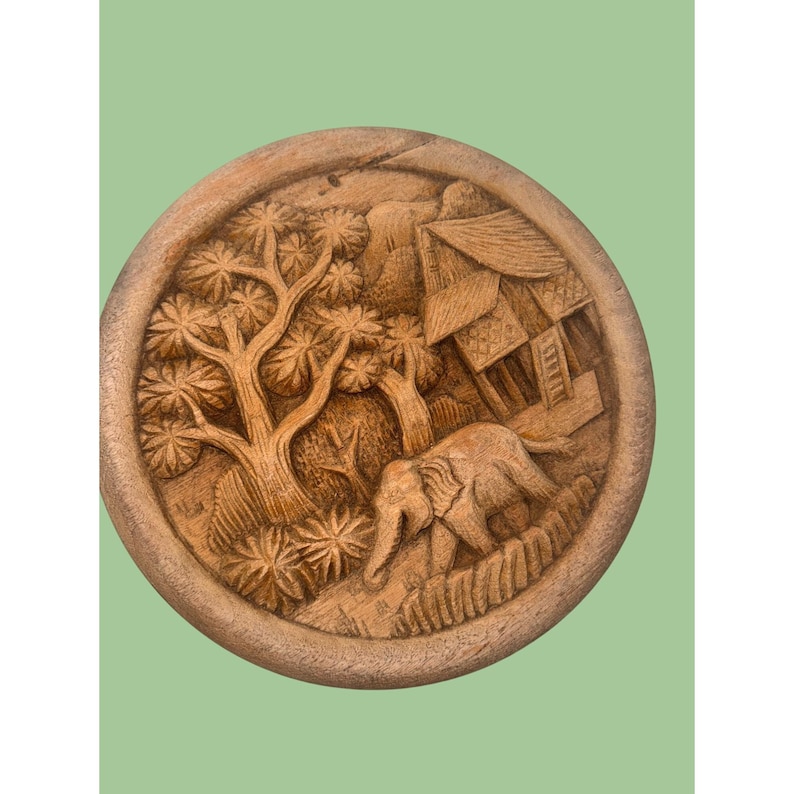 May include: A round, wooden carving depicting a detailed landscape scene. The carving features a tree, a building, and an elephant walking on a path. The artwork is in shades of brown and has a rustic, handcrafted appearance.