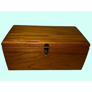 May include: A rectangular wooden box with a hinged lid and a metal clasp. The box is made of light brown wood with a visible wood grain pattern. The clasp is dark metal, and the box appears to be empty.