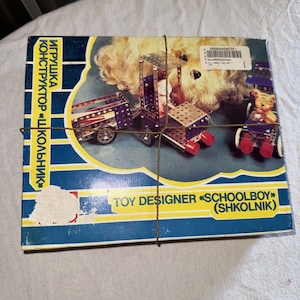 Może przedstawiać: Niebiesko-żółte pudełko z napisem "Игрушка Конструктор "Школьник" Toy Designer "Schoolboy" (Shkolnik)" z przodu. Pudełko zawiera metalowy zestaw konstrukcyjny z ciężarówką, przyczepą i małym samochodem.