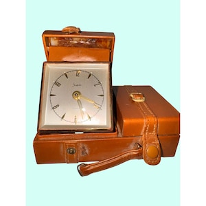 Könnte beinhalten: Eine Vintage-Reiseuhr mit quadratischem Zifferblatt und braunem Lederetui. Das Zifferblatt ist silberfarben mit schwarzen Ziffern und goldenen Zeigern. Das Etui ist geöffnet und zeigt die Uhr, daneben ein kleineres Etui.
