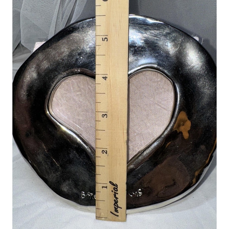 May include: A silver-colored heart-shaped picture frame with a light pink interior. The frame is approximately 5 inches tall, as measured by a wooden ruler. The frame has a unique, irregular shape.