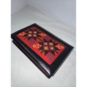 May include: A rectangular black box with a red embroidered top featuring a geometric pattern in shades of yellow, blue, and brown. The box has a black frame and appears to be closed.