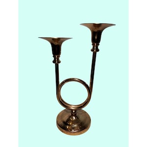 Puede incluir: Un candelabro de latón con dos copas acampanadas. El candelabro tiene una base circular y un diseño curvo en forma de cuerno que conecta las copas. El metal tiene un tono dorado cálido.