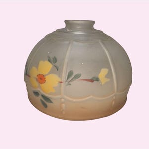 Może przedstawiać: Matowy szklany klosz lampy o zaokrąglonym kształcie i falistym brzegu. Klosz posiada ręcznie malowany kwiatowy wzór z żółtymi kwiatami, pomarańczowymi środkami i zielonymi liśćmi. Podstawa klosza ma miękki, kremowy kolor.