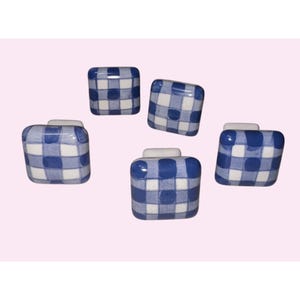 May include: Five square cabinet knobs with a blue and white gingham pattern. Each knob has a white base and a rounded, rectangular shape. The knobs are arranged against a light pink background.