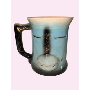 Puede incluir: Una taza de cerámica con un diseño de cara único. La taza presenta un cuerpo azul claro con un asa negra y dorada. El diseño de la cara incluye una boca redonda y blanca y dos líneas horizontales para los ojos. El borde superior es de color beige.