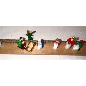 Op de afbeelding: Een set miniatuur kerststalfiguren, waaronder een engel, een kribbe en verschillende figuren in gekleurde gewaden, gerangschikt langs een houten liniaal die laat zien dat de stukken ongeveer 2,5 tot 5 cm hoog zijn. De figuren zijn geschilderd in groen, rood, blauw en wit.