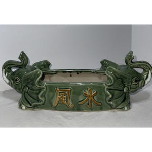 May include: Green ceramic planter with two elephant head sculptures on each end. The planter has a rectangular shape with the Chinese characters "Feng Shui" in gold lettering on the front. The elephants have white tusks and detailed features.