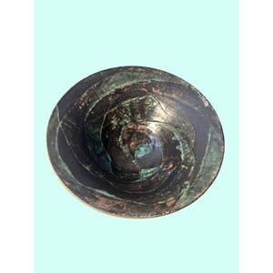 Puede incluir: Un cuenco redondo con una superficie oscura y desgastada que presenta patrones en espiral de negro profundo y verde azulado oxidado. El interior muestra una apariencia texturizada y estratificada con tonos metálicos bajo el acabado.