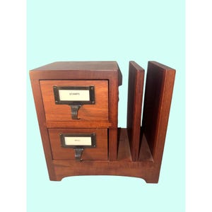 Puede incluir: Un organizador de escritorio de madera marrón oscuro con dos cajones apilados con tiradores y portaetiquetas de metal. El cajón superior tiene la etiqueta STAMPS y el inferior MISC. A la derecha de los cajones hay dos ranuras verticales.