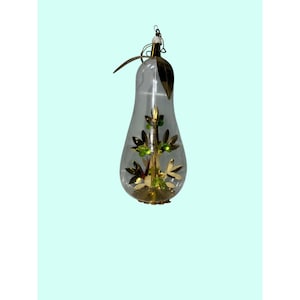 May include: Clear glass Christmas ornament shaped like a pear, with gold and green accents. The ornament has a gold base and a gold leaf at the top. Inside is a gold tree with green jewels.