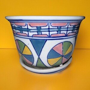 May include: A white ceramic planter with a geometric design in blue, green, pink, and yellow. The planter has a matching saucer.
