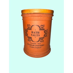 May include: A cylindrical, orange-brown Fulham Pottery bath salts jar with a wooden lid. The jar features black text that reads "BATH SALTS" and "Founded 1671 Fulham Pottery London, England."