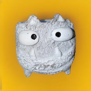 Peut inclure: Un pot en céramique blanche en forme de monstre avec de grands yeux et un sourire narquois. Le pot a une surface texturée et est parfait pour ajouter une touche fantaisiste à n'importe quelle maison.
