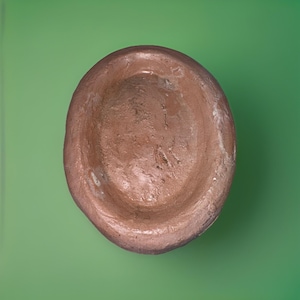 May include: A handmade, oval-shaped, brown ceramic bowl. The bowl has a rough, textured surface and a slightly uneven rim. The interior is a darker shade of brown than the exterior. The bowl is set against a green background.