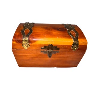 Può includere: Un piccolo forziere in legno con la parte superiore curva e ferramenta in ottone. Il forziere è realizzato in legno marrone-rossastro e presenta cerniere e chiusura decorative in metallo. Il design generale è vintage e rustico.