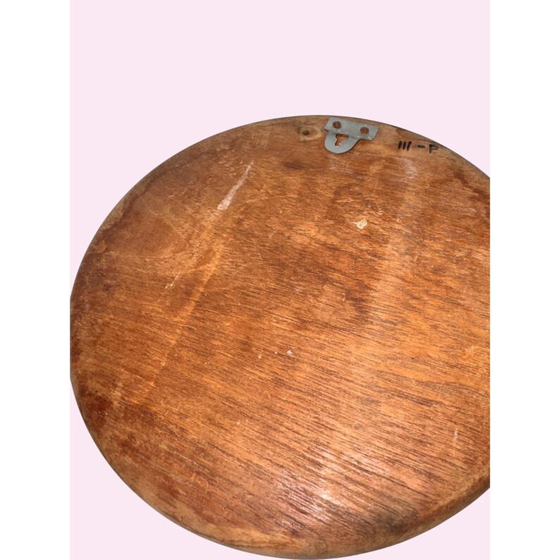 May include: A round, wooden decorative piece with a natural wood grain finish. It features a metal hanging bracket on the top. The piece has a slightly irregular shape and a warm, brown color.