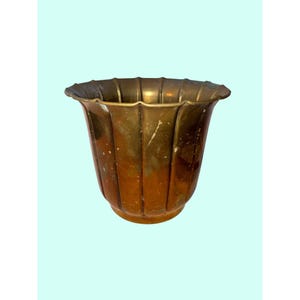 May include: A fluted brass planter with a wide, scalloped rim and a tapered base. The metal surface displays a warm, aged patina with visible variations in color, ranging from deep bronze tones to lighter, metallic gold highlights throughout.