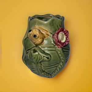 Puede incluir: Un macetero de pared de cerámica verde con forma de hoja con una rana y una flor.