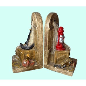 May include: Pair of nautical-themed bookends. Each bookend features a weathered wood-look design with a different nautical element: an anchor and a red lantern. Additional details include rope, a chest, and a buoy.
