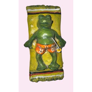 May include: A green frog figurine sunbathing on a yellow towel with black, red, and white stripes. The frog wears orange swim trunks with white flower patterns and holds a small, round object.