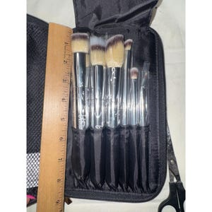 Puede incluir: Un juego de siete brochas de maquillaje con mangos plateados y cerdas de puntas blancas, organizadas en un estuche negro con cremallera. Una regla de madera está colocada junto a las brochas, mostrando una longitud de unos 18 cm.