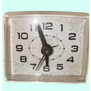 May include: A vintage Sunbeam alarm clock with a clear plastic case. The clock face is white with black numerals and gold-colored hands. The face has a decorative floral pattern and the word "Sunbeam".
