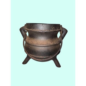 May include: A dark brown cast iron cauldron with a rounded body, three short legs, and two side handles. A thin, curved metal handle is attached to the side lugs, arching over the top of the vessel. The surface shows a textured, weathered finish.