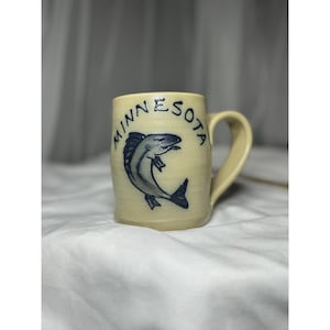 May include: A cream-coloured ceramic mug with a curved handle. The mug features a blue illustration of a fish and the word "MINNESOTA" in a circular design around the top. The mug is on a white surface.