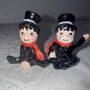 May include: Two vintage ceramic figurines of chimney sweeps. Each figure is dressed in black with a red scarf and a black top hat. The figurines have painted facial features and are posed in a seated position.
