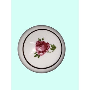 Op de afbeelding: Witte keramische kastknop met een geschilderde roze roos en groene bladeren. De ronde knop heeft een zwarte rand. Deze decoratieve knop kan worden gebruikt voor laden of kasten.
