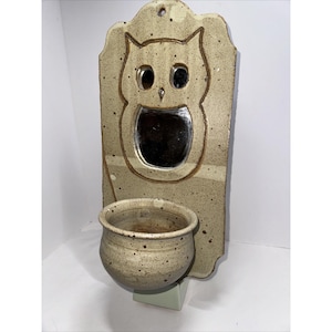 May include: A ceramic wall planter featuring an owl design with a small mirror in the owl's face. The planter has a speckled brown and white glaze and a small, round pot attached to the bottom.