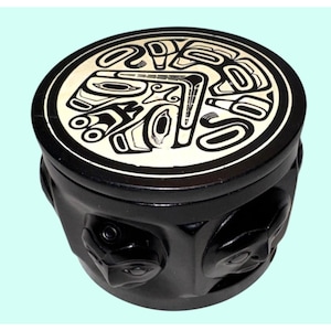 May include: A round black lidded box featuring a cream-colored circular top with a black graphic line art design. The sides of the box are black and decorated with multiple raised, sculpted faces that wrap around the exterior of the container.